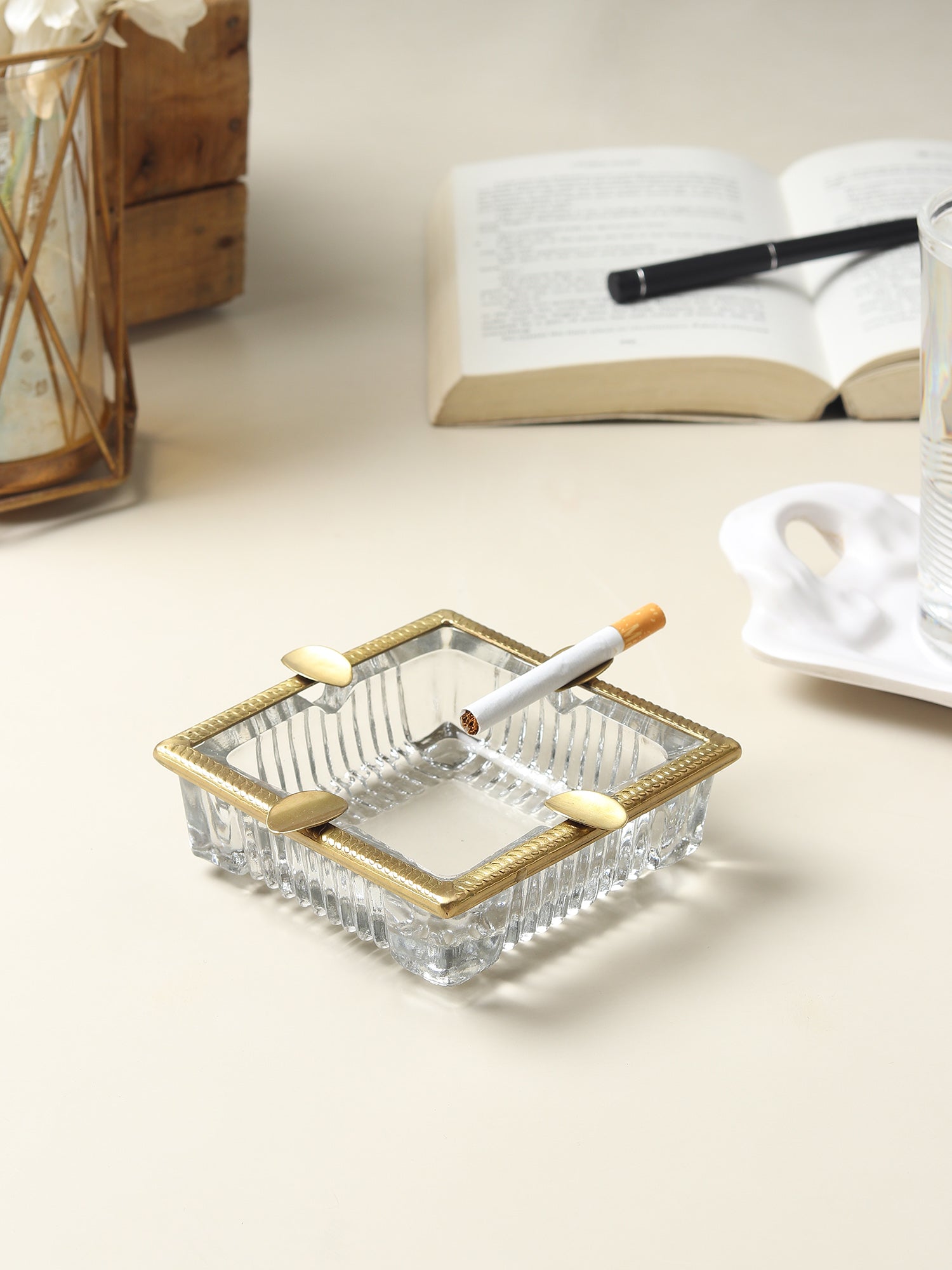 Glass ashtray with gold rim on a light surface with a book and decorative items in the background.