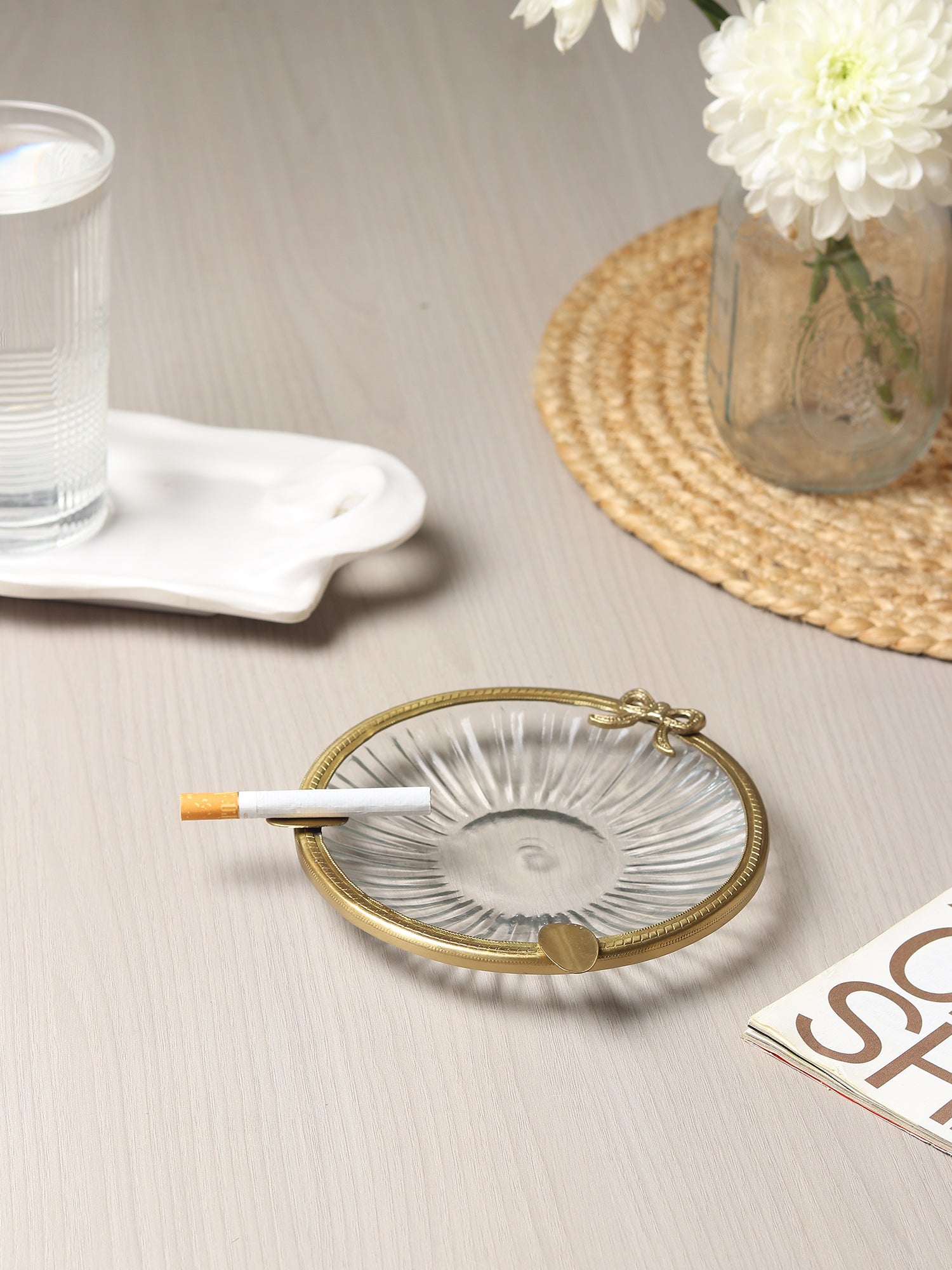 Glass ashtray with gold rim on a table with a cigarette, glass of water, and flowers.