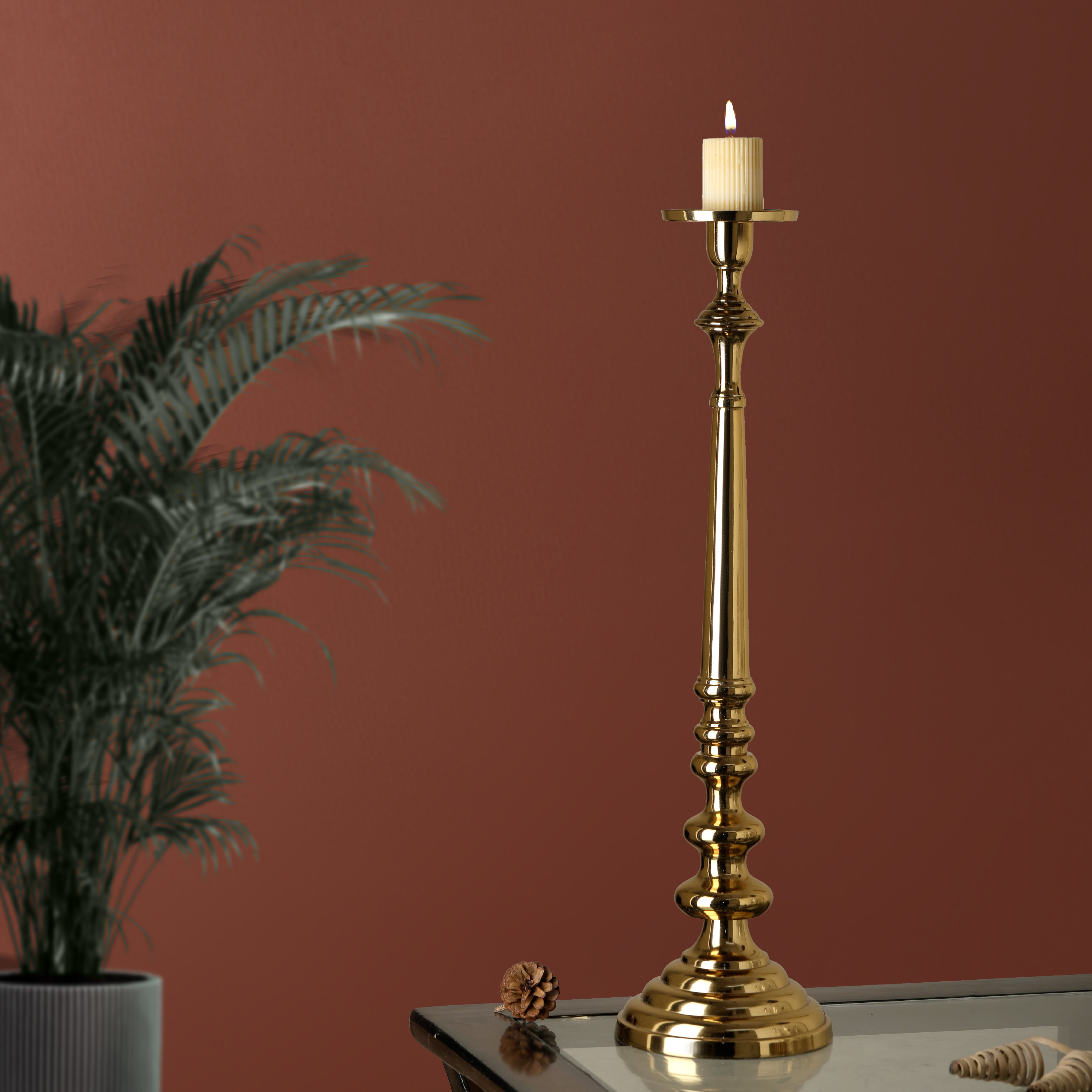 Gold candlestick with a candle on a surface against a red wall with a plant to the left.